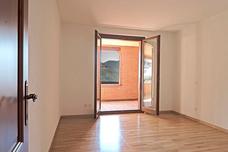 Spacious room with hardwood flooring, white walls, and a large window with a brick frame providing a view of the mountains outside.