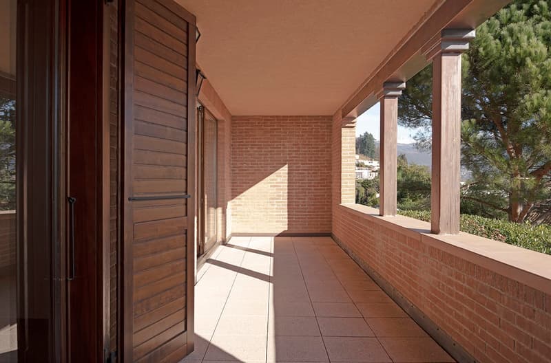 Covered porch with brick walls, wooden beams, and tiled flooring. Large windows provide a view of the surrounding trees and landscape.