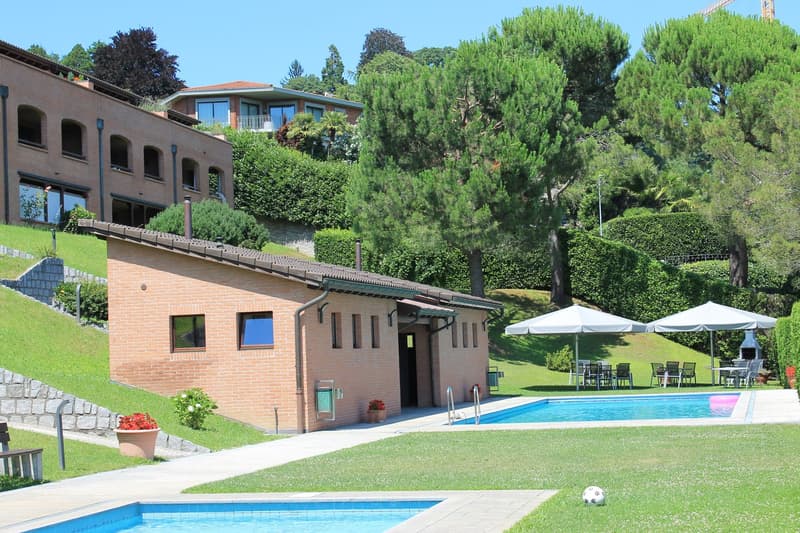 Two-story brick building with large windows, surrounded by lush greenery and trees. There is a swimming pool in the foreground, and a patio area with umbrellas and tables. The building appears to have balconies or terraces on the upper level.