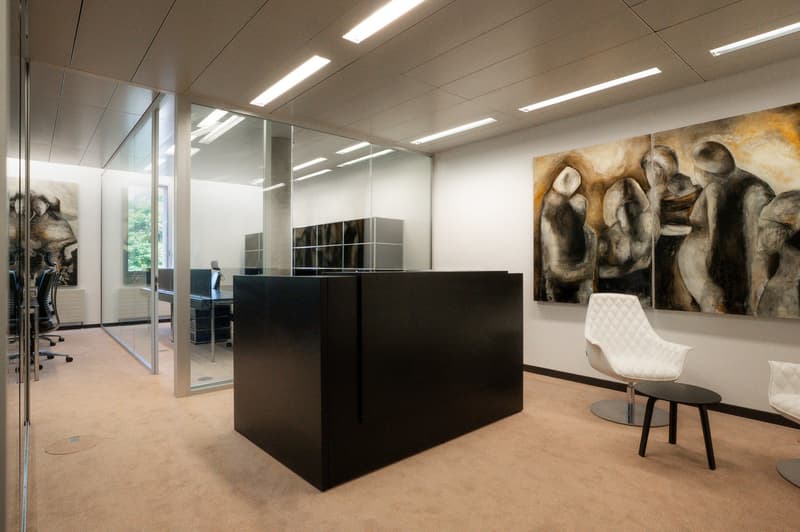 Modern and minimalist office space with glass walls, black furniture, and a large abstract painting on the wall. The space has recessed lighting and a comfortable-looking white chair.