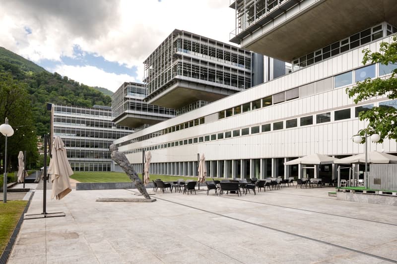 Multi-story modern office building with glass and concrete facade, outdoor seating area with umbrellas, surrounded by greenery and mountains in the background.