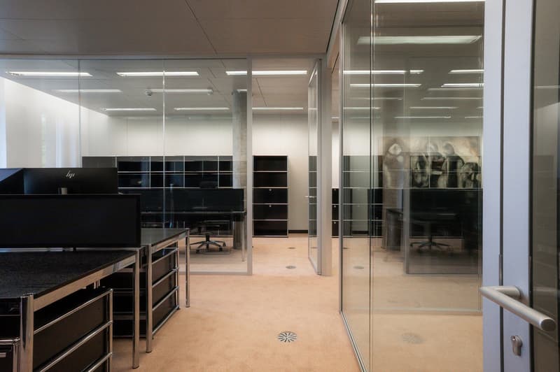 Open office space with glass walls, desks, computers, and storage units. The space appears to be well-lit with overhead lighting and has a modern, minimalist design.