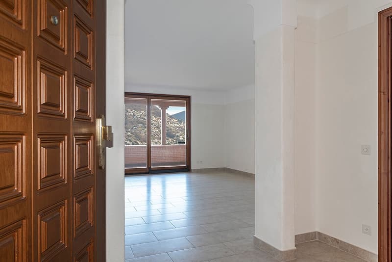 Large wooden entrance door, white walls, tiled floor, and a window with a view of the mountains.