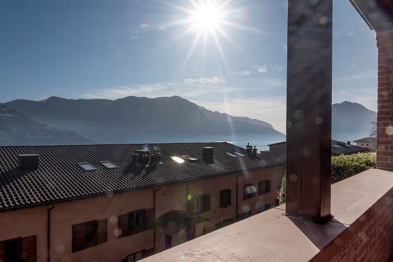 The image shows a building with a tiled roof overlooking a scenic mountain landscape. The sun is shining brightly in the sky, creating a warm and inviting atmosphere. The building appears to be situated on a hillside, offering a panoramic view of the surr