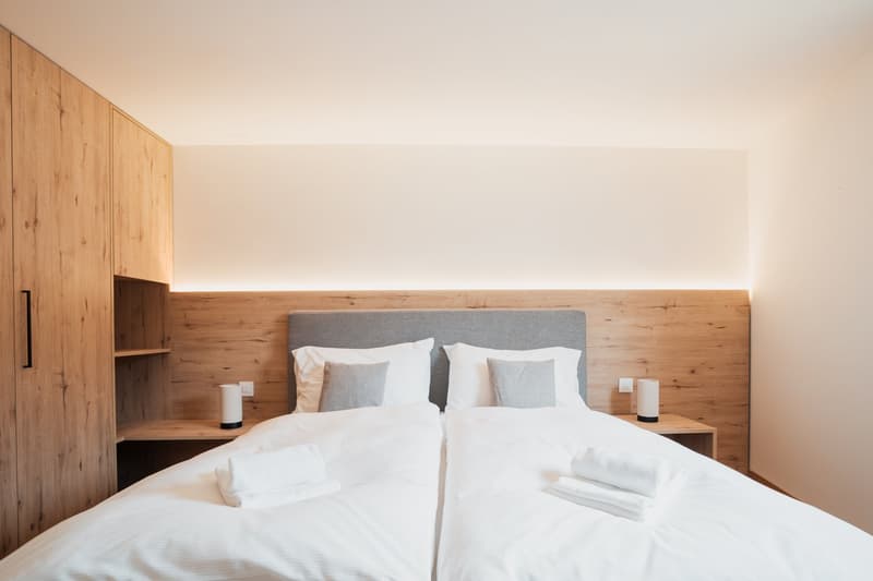 Twin bed, white bedding, wooden walls, bedside tables, lamps, two nightstands, two towels on each bed, wooden shelves, and an electrical outlet.