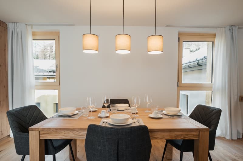 Dining room with white walls, wooden table, four chairs, pendant lights, and two windows with curtains