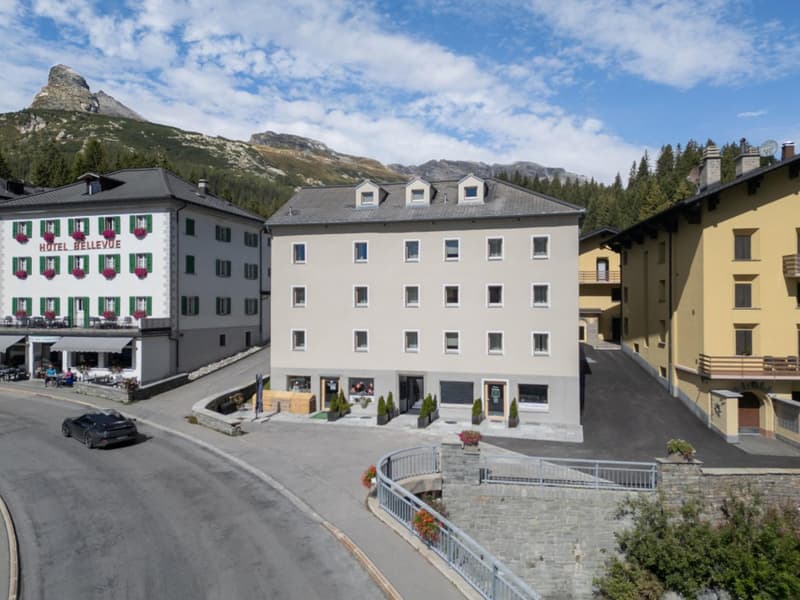 Three-story hotel building, several windows, balconies on the upper floors, mountain view, parked car, paved road, stone fencing.