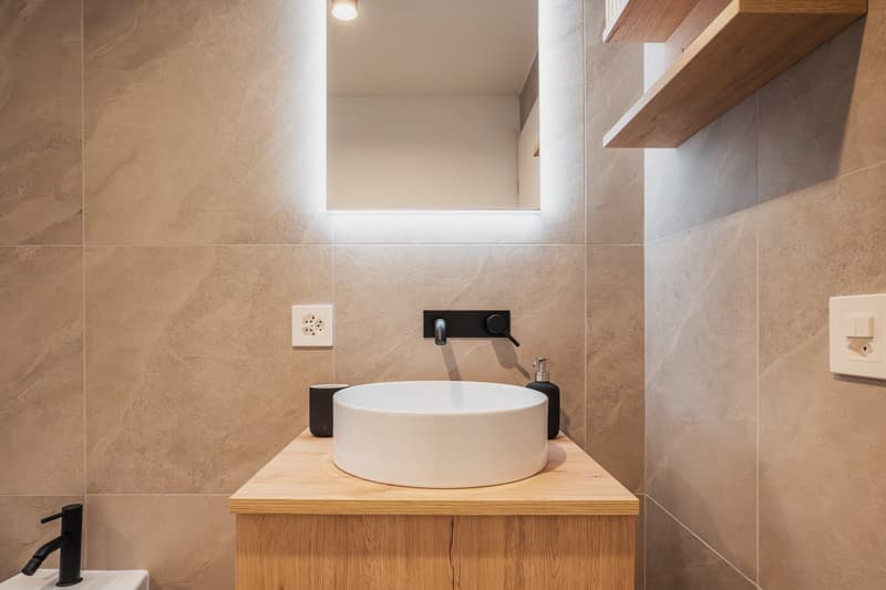 Bathroom with white sink, brown countertop, dark brown tiles, small wall mirror, wooden shelves, and black taps and faucets.