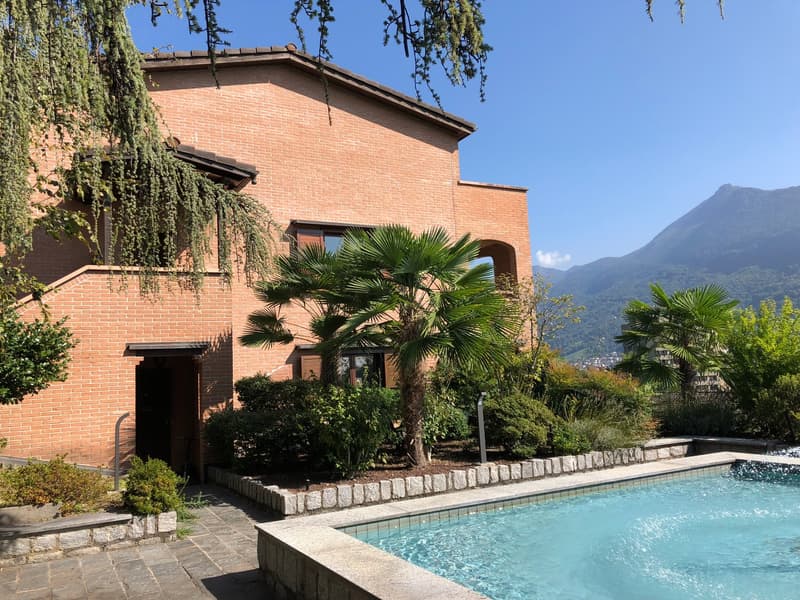 two-story brick house, rectangular swimming pool, lush greenery, wooden door, tiled walkway, mountains in the background
