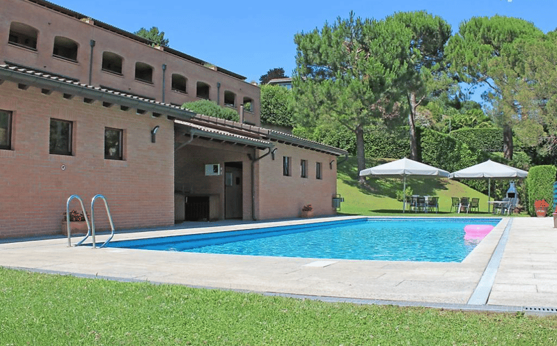 Building exterior with swimming pool, brick walls, tiled roof, outdoor seating, umbrellas, potted plants, green lawn, trees.