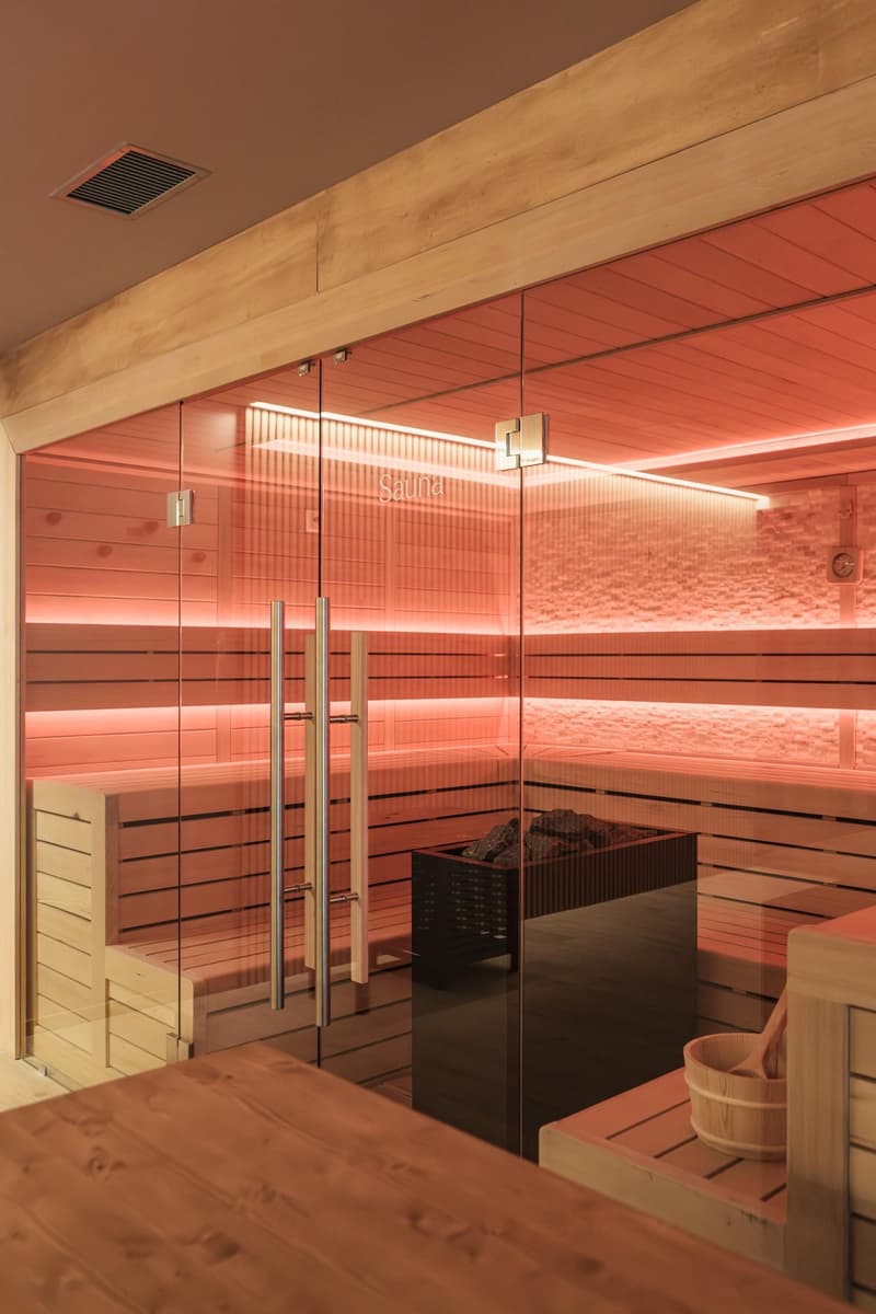 A sauna room with wooden benches, a wooden bucket, and a wooden spoon. The room has a glass door with a silver handle and a wooden wall. The sauna heater is on the right side of the room, and the word 'Sauna' is written on the glass door. The room is illu