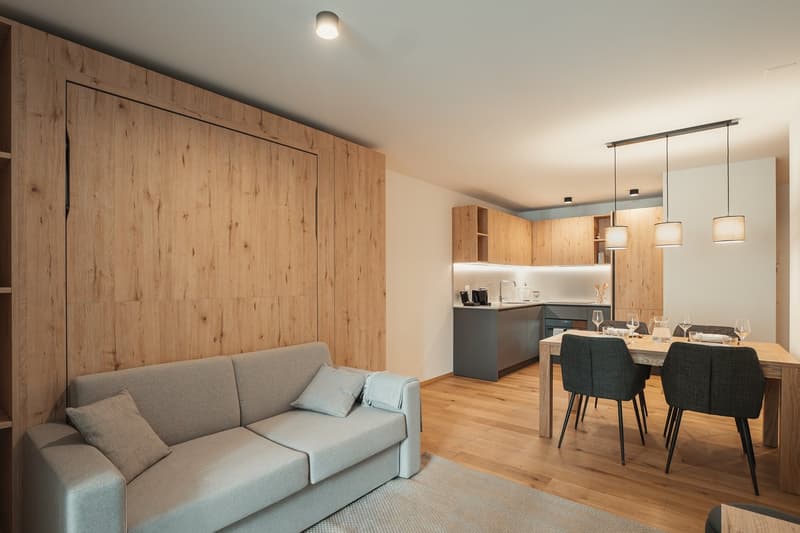 Open-plan living and dining area with a gray sofa, dining table, and wooden flooring. The kitchen is partially visible with a sink and cabinets.