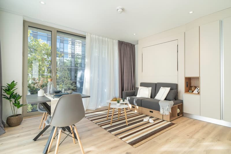 Spacious living room with large windows, gray sofa, wooden coffee table, striped rug, and a dining area with a table and chairs. The room has a modern and minimalist design with neutral colors and natural materials.