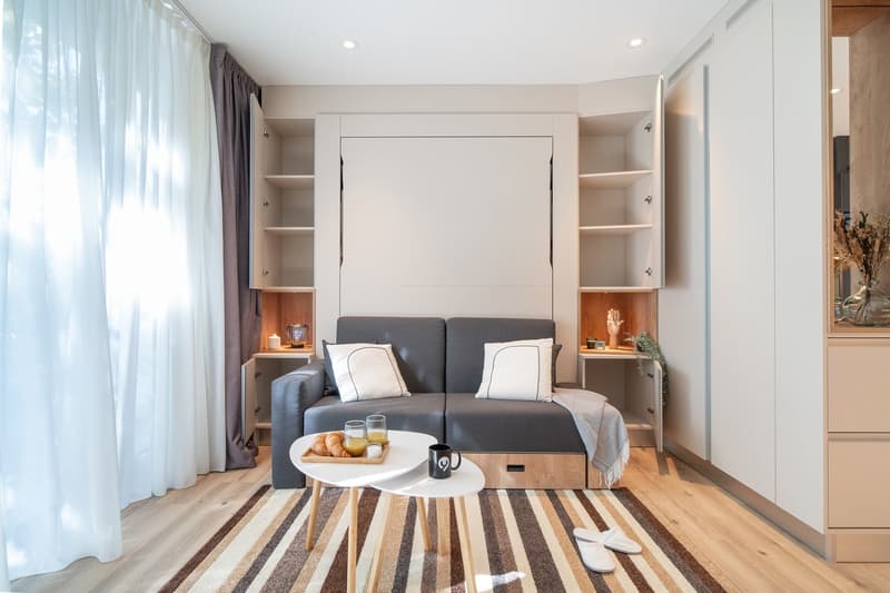 The image shows a modern and cozy studio apartment. The room features a gray sofa bed with white pillows, a round white coffee table, and built-in shelving units on either side of the bed. The room has large windows with gray curtains, providing natural l