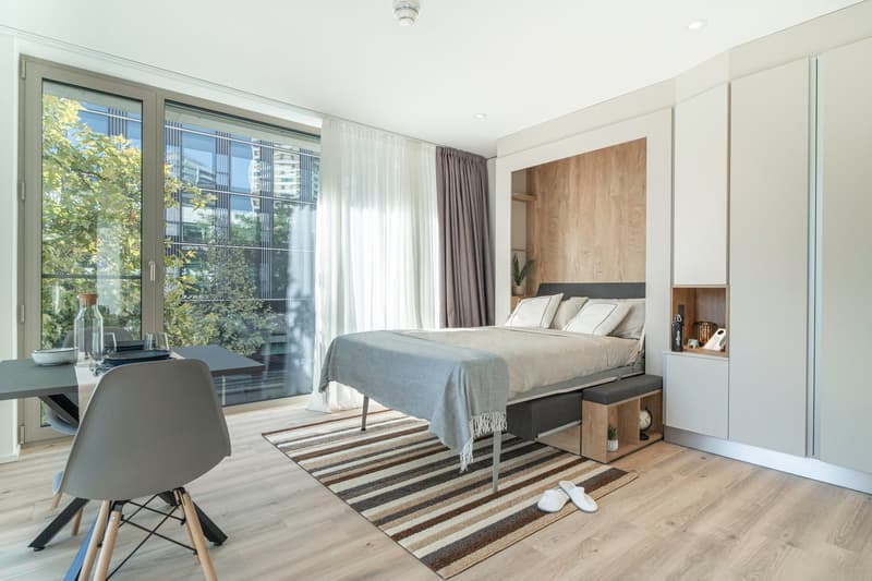 Spacious bedroom with large windows overlooking a balcony, hardwood flooring, a king-size bed with gray bedding, a wooden accent wall, built-in shelving, and a striped area rug.