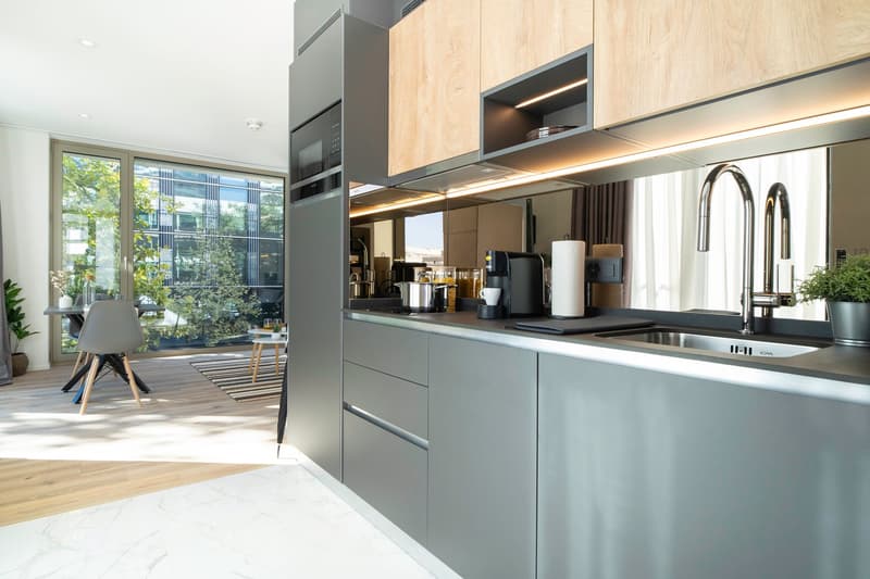 Modern kitchen with gray cabinets, wooden accents, built-in appliances including a dishwasher, and large windows overlooking a garden area.