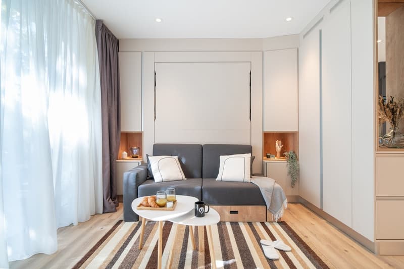Spacious living room with large windows, gray sofa, coffee table, wooden side tables, and a striped rug on the hardwood floor.
