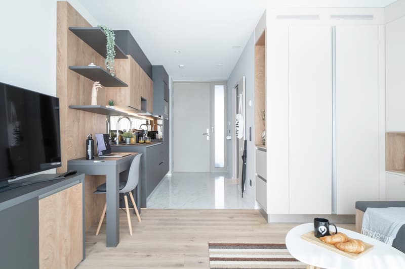 The image shows a modern and minimalist studio apartment. The space features a kitchen area with gray cabinets, open shelves, and a small dining table. There is a TV and entertainment unit in the living area, and the apartment has a bright and airy feel w