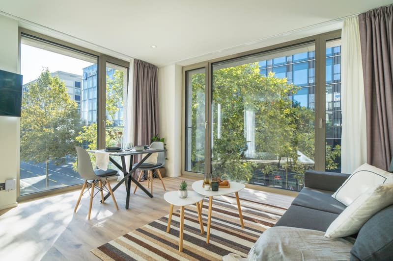 Spacious living room with large windows overlooking a balcony and a view of the city. The room features a dining table, chairs, coffee table, and a sofa. The decor is modern and minimalist, with a striped rug on the hardwood floor.