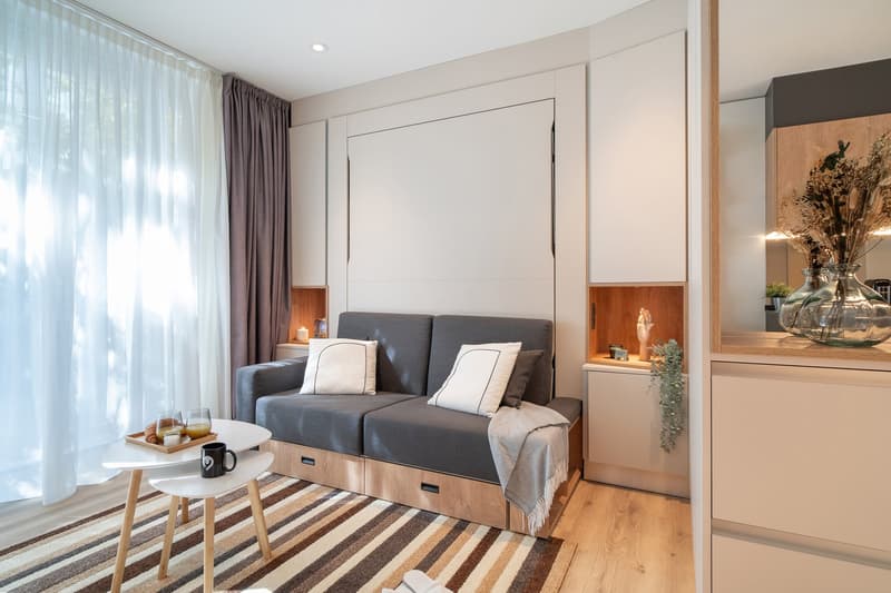 Cozy living room with a gray sofa, wooden storage units, and a striped rug. Large windows provide natural light and access to a balcony or terrace.
