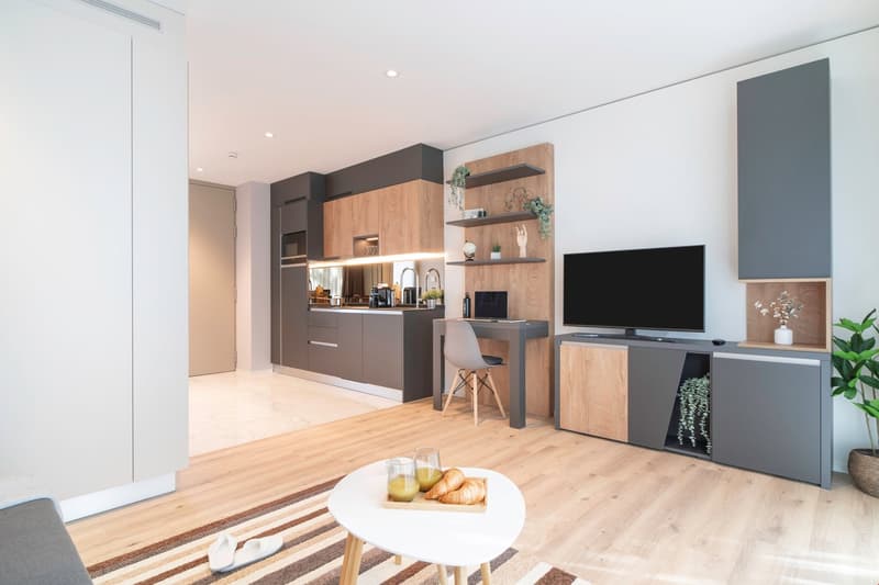 Open-concept studio apartment with modern kitchen, living area, and workspace. The kitchen features dark cabinets and wood accents, with a dishwasher and washing machine visible. The living area has a TV mounted on the wall, and a round coffee table with
