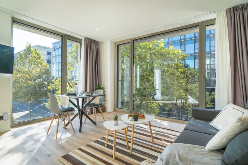 Spacious living room with large windows overlooking a balcony and a view of the surrounding greenery. The room features a dining table, chairs, coffee table, and a sofa. The decor has a modern and minimalist style with wooden and neutral-toned elements.