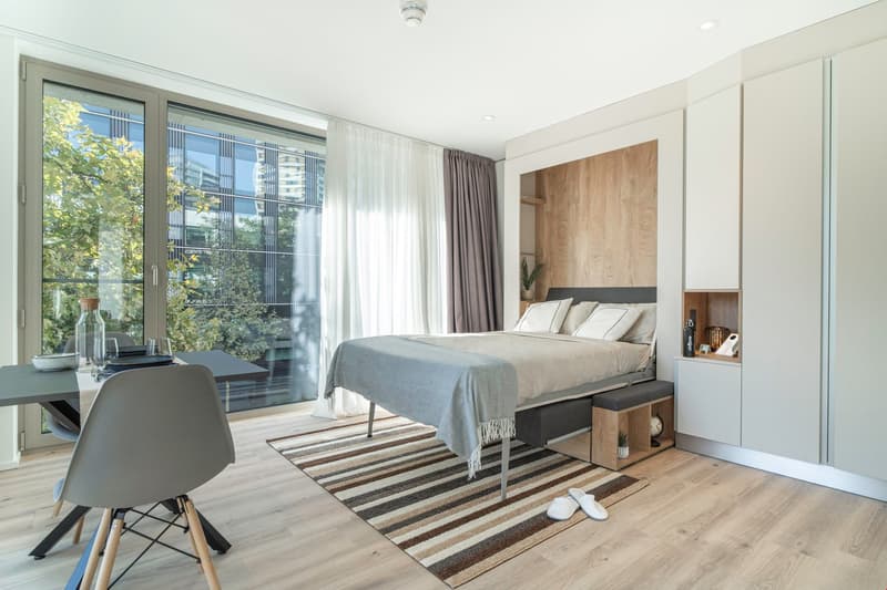 Spacious bedroom with large windows overlooking a balcony, hardwood flooring, a striped area rug, a modern bed frame with built-in storage, and a wooden accent wall with shelving. The room also features curtains, a desk, and a chair.