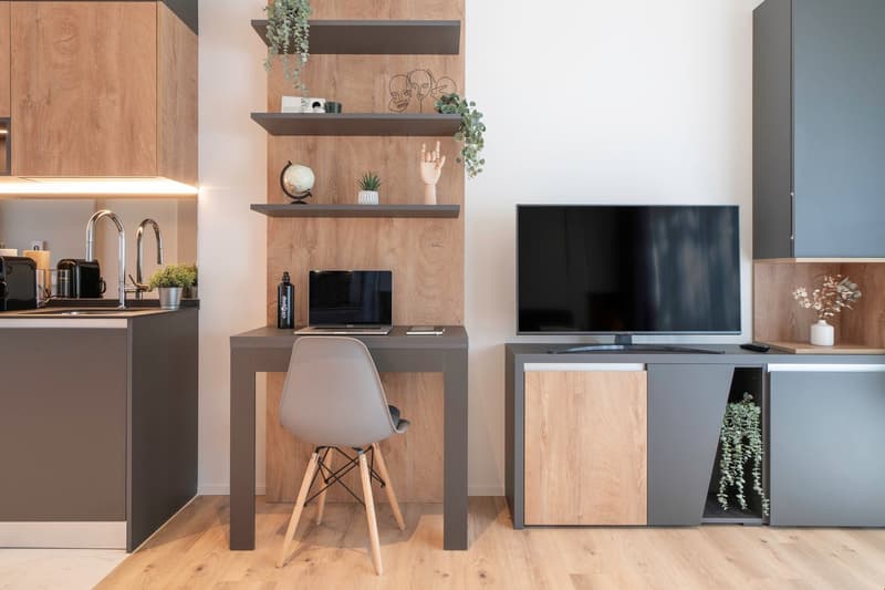 Modern studio apartment with open kitchen, dining and living area. Features include wooden shelves, TV stand, laptop desk, and a white chair. The kitchen has a dishwasher and washing machine visible.