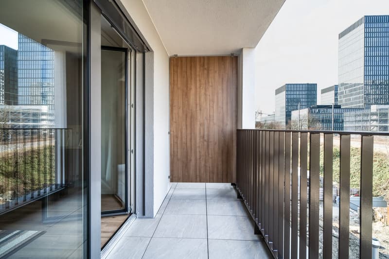 Spacious balcony with sliding glass doors, wooden wall paneling, and a tiled floor. The balcony overlooks a cityscape with modern high-rise buildings in the background.