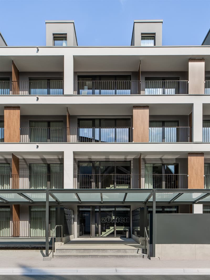 A modern multi-story building with a glass and concrete facade. The building has multiple balconies on each floor, with wooden accents. The main entrance is located at the ground level, with a glass and metal canopy above it.