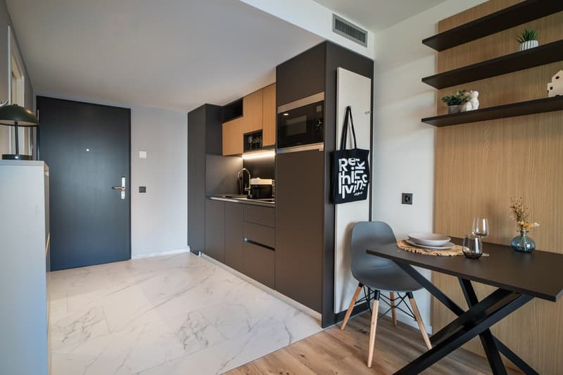 The image shows a modern and stylish studio apartment. The space features a kitchen area with dark cabinets, a built-in microwave, and a dishwasher. There is a dining table with chairs in the living area, and the flooring is a mix of marble tiles and hard