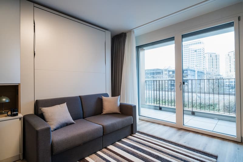 Modern living room with large windows, gray sofa, striped rug, and access to a balcony or terrace with a view of the city skyline.