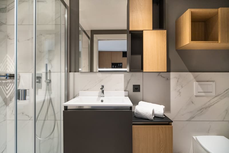 Modern bathroom with marble-like tiles, glass shower enclosure, vanity with sink, wooden storage cabinets, and towels