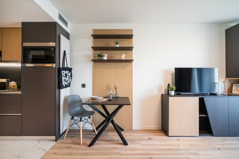 Modern studio apartment with open kitchen, dining area, and living room. The kitchen features dark cabinets and a built-in microwave. The dining area has a small table with a single chair. The living room has a TV stand and shelves displaying decor items.