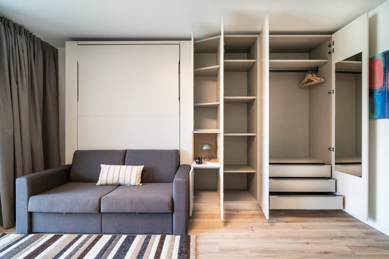 Compact studio apartment with a gray sofa, built-in storage cabinets and shelves, and a raised sleeping area with steps leading up to it. The room has hardwood floors and large windows, creating a bright and airy atmosphere.