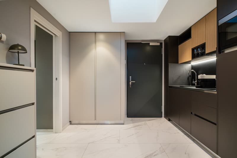 Compact studio apartment with open-plan layout. Marble-like tile flooring, gray walls, built-in kitchen with dark cabinets and a dishwasher, and a sliding door leading to the bathroom. The entrance door is visible, along with a small refrigerator and a la