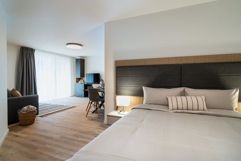 Spacious bedroom with hardwood flooring, large windows, a king-size bed with a padded headboard, and a seating area with a sofa and desk. The room also has a balcony or terrace visible.