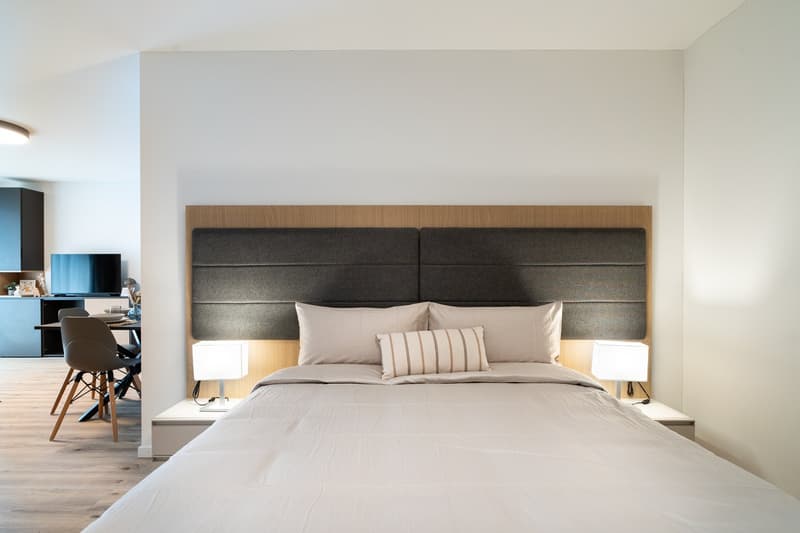 Spacious bedroom with a large bed, wooden headboard, and bedside tables with lamps. The room has a minimalist and modern design with white walls and hardwood flooring.