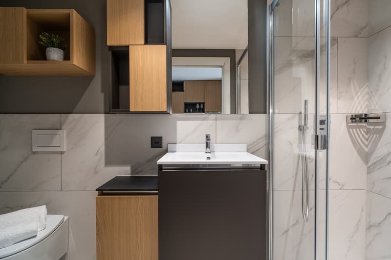 Modern bathroom with marble-like tiles, wooden vanity cabinet, white sink, and glass shower enclosure