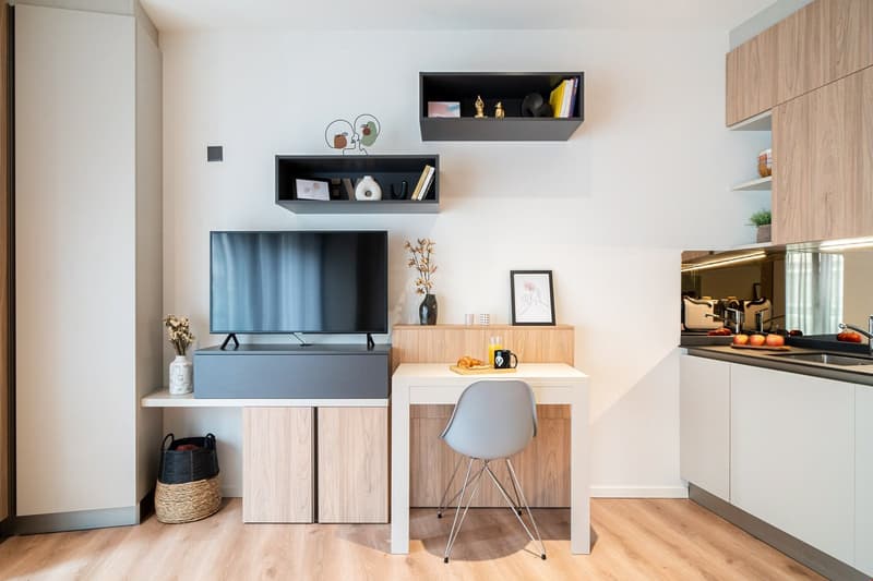 The image shows a modern and minimalist living room with a TV stand, shelves, and a small desk area. The TV stand has a black TV mounted on it, with storage cabinets below. The shelves above the TV stand display various decorative items. The desk area has