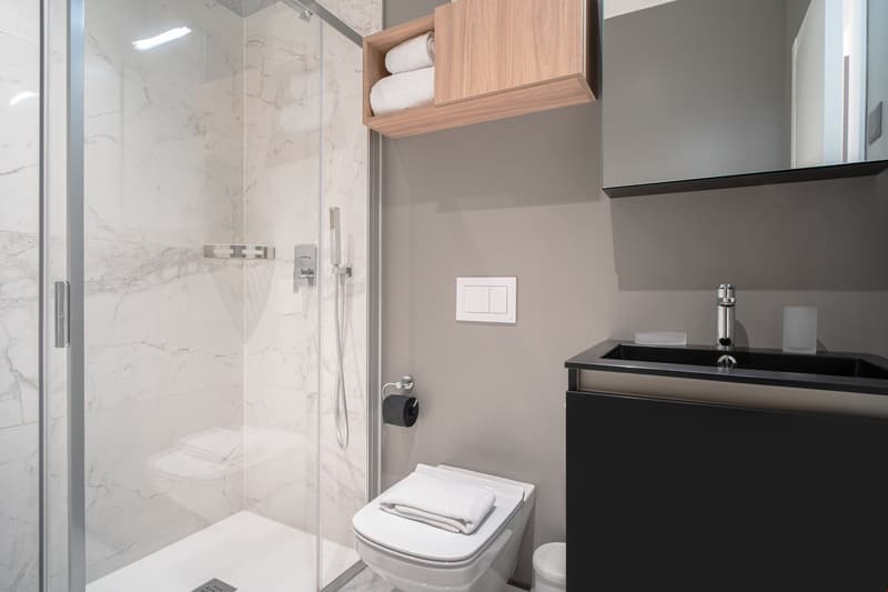 Modern bathroom with marble-like tiles, glass shower enclosure, vanity with black countertop, and a built-in storage cabinet above the toilet.