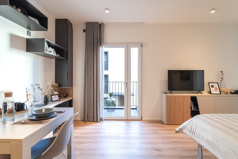 Open-plan studio apartment with kitchenette, living area, and bedroom. Features include hardwood flooring, large windows with balcony access, built-in shelving, TV stand, and a desk area.