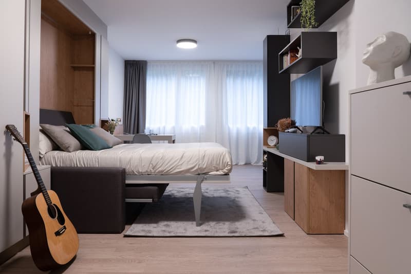 Spacious bedroom with large windows, hardwood flooring, modern furniture including a bed, dresser, and shelving unit, and a guitar in the corner.
