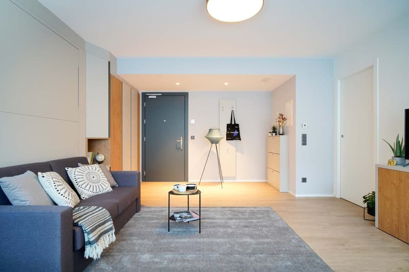 Modern living room with gray sofa, decorative pillows, coffee table, and a floor lamp. The room has light-colored walls, hardwood floors, and a balcony or terrace visible through the doorway.