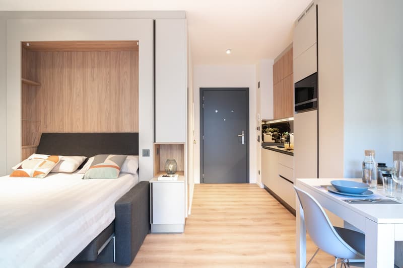 The image shows a modern studio apartment with a bedroom area, a kitchenette, and an entryway. The bedroom features a wooden headboard and built-in storage. The kitchenette has a dishwasher and other appliances. The entryway has a dark gray door leading t