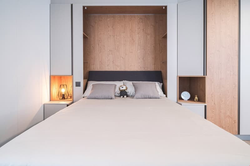 Spacious bedroom with wooden paneled walls, a large bed with gray bedding, and a built-in wardrobe with lighting. There is also a small shelf unit next to the bed.