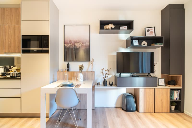 The image shows a modern and stylish open-plan living room and kitchen area. The kitchen features wooden cabinets and a built-in microwave. The living room has a TV stand with shelves, a framed artwork, and decorative items. The dining area has a white ta