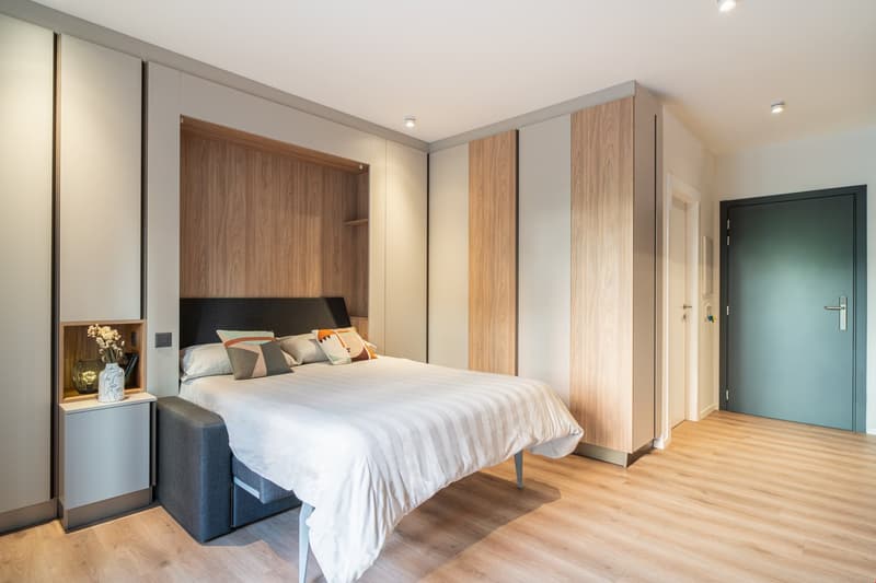 Spacious bedroom with wooden flooring, gray and white color scheme, large bed with headboard, built-in wardrobe, and bedside tables. The room also features a balcony or terrace area.