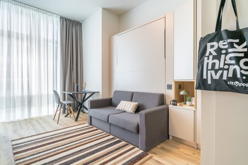 Spacious living room with large windows, gray sofa, striped rug, dining table, and a large tote bag with text on it.