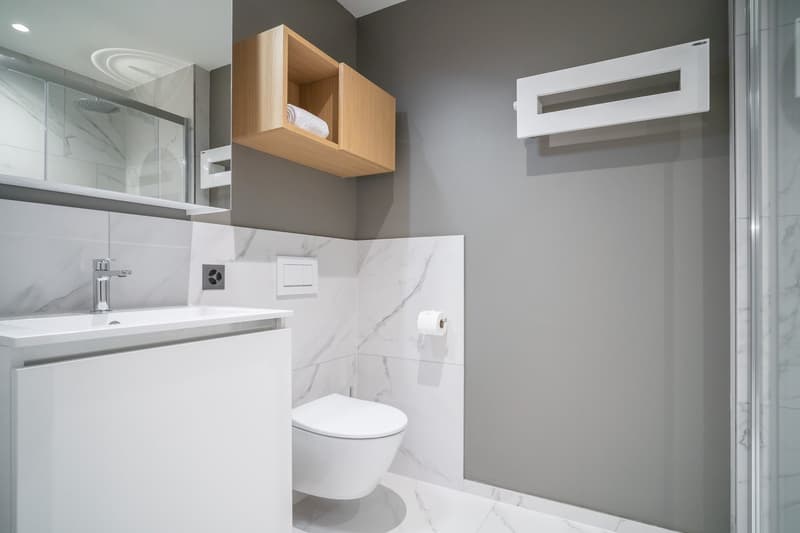 Modern bathroom with white marble-like tiles, a vanity with a sink, a toilet, and a wooden storage cabinet above the toilet. The bathroom also features a towel rack and a recessed lighting fixture.